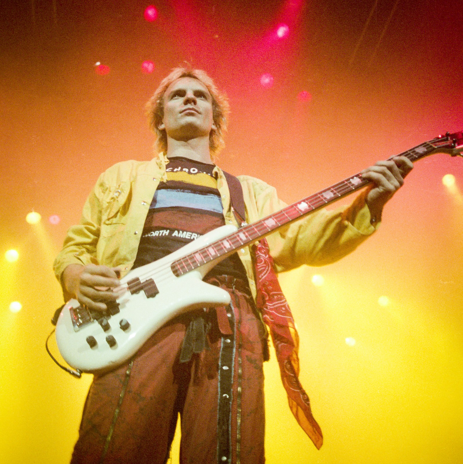 Sting of The Police playing a white bass under red and yellow stage lights at Buffalo Memorial Auditorium, 1984 Synchronicity Tour - concert photography by Chuck Spang